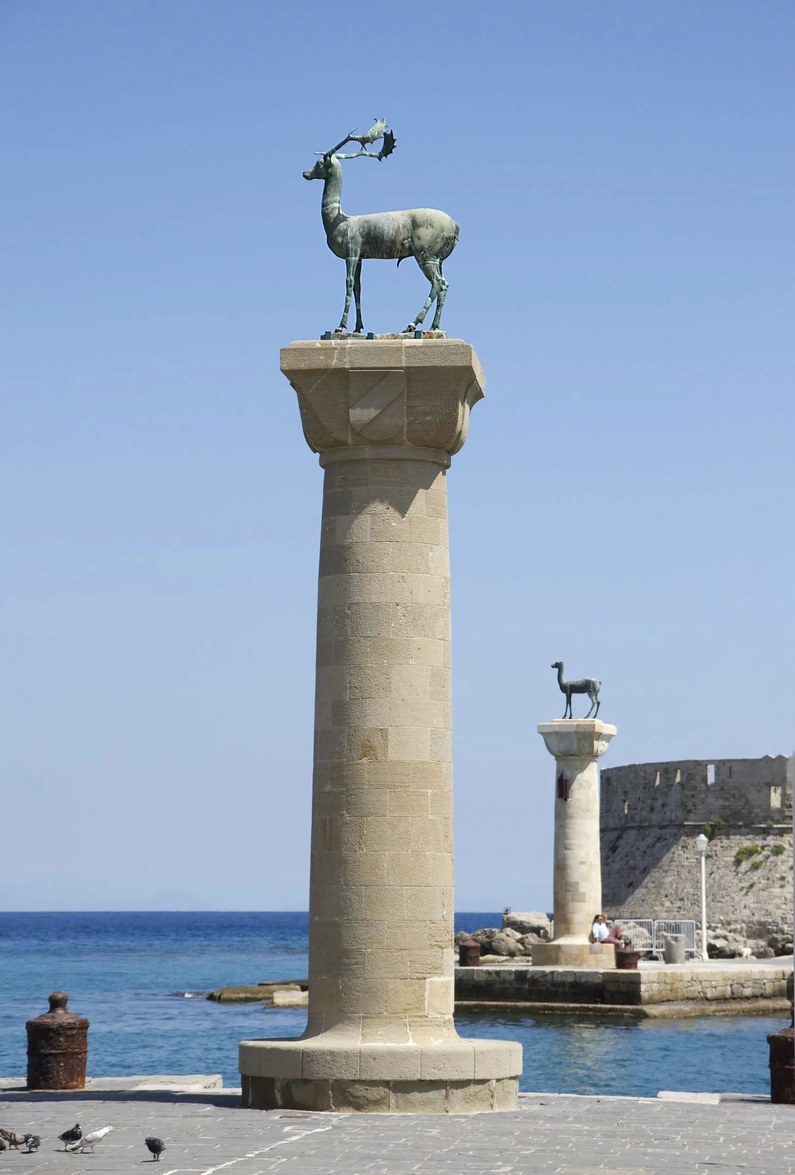 Zwei Säulen mit Hirschstatuen an der Einfahrt zum Mandraki-Hafen, im Hintergrund eine Festung